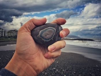 Close-up of hand holding pebble at beach against cloudy sky