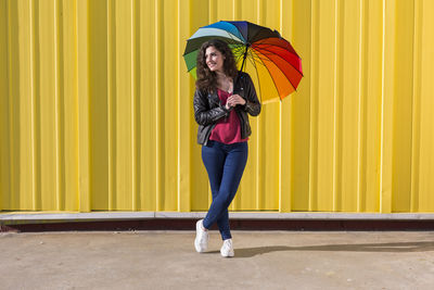Full length of smiling mid adult woman holding colorful umbrella while standing against yellow wall
