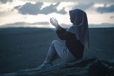 Woman holding illuminated string lights while sitting on tree trunk during sunset