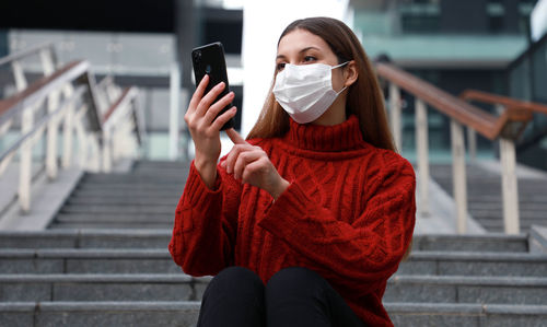 Low angle view woman wearing mask using smart phone while standing outdoors