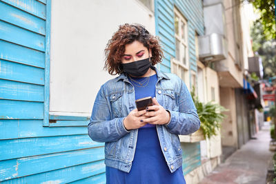 Plus size female wearing protective mask standing in narrow street near residential building and browsing smartphone during covid 19 epidemic in city