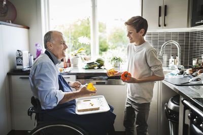 Happy disabled father in wheelchair preparing food with son in kitchen