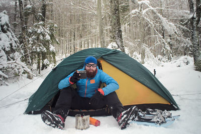 Portrait of young man with coffee sitting in tent on snow covered forest