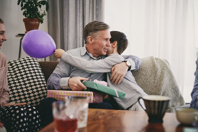 Grandfather holding birthday gift while embracing grandson on sofa at home
