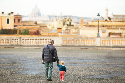 Rear view of grandfather holding granddaughter hand while walking on road