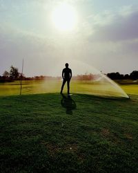 Silhouette man standing on grassy field by agricultural sprinkler during sunny day