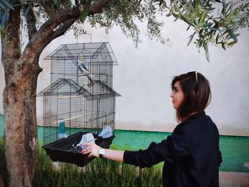 Side view of young woman looking at bird in cage while standing against wall