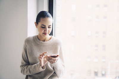 Woman using mobile phone while standing by window at home