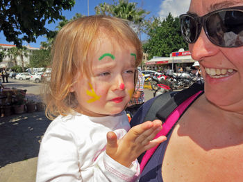 Mother carrying girl with painted face crying against trees and sky