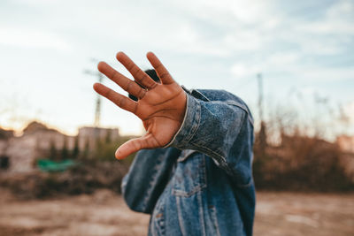 Anonymous ethnic male in denim jacket hiding face with reached arm under cloudy sky in daylight