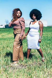 Full body plus size african female models in trendy outfits with curly hair standing on green grass against cloudless blue sky on sunny day in meadow