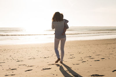 Rear view of mother with baby walking at beach against clear sky during sunset