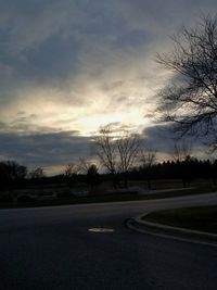 Road by bare trees on field against sky at sunset