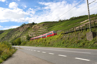 A red passenger train driving alongside an empty motorway next to the hills of a vineyard in germany