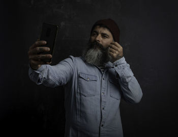 Man with beard making a selfie sticking his mustache. jeans shirt, wool hat and mobile in hand
