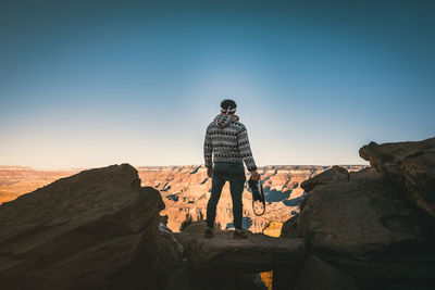 Rear view of man holding camera while standing on rock against sky