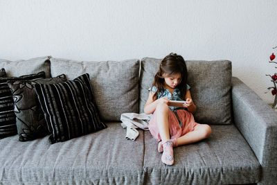 Full body of adorable ethnic little girl in casual clothes and socks relaxing on comfortable sofa and playing video game with smartphone