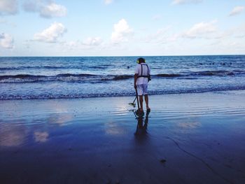 Full length rear view of man holding metal detector on beach