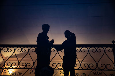 Silhouette couple standing on railing against sky during sunset