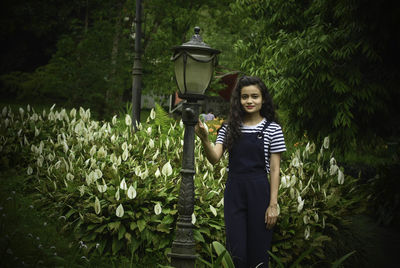 Portrait of young woman standing by street light against trees in park