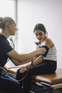 Female nurse checking blood pressure of girl sitting on bed in clinic