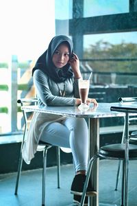 Full length of woman wearing hijab looking away while sitting at cafe
