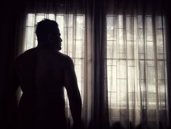 Rear view of silhouette man looking through window at home