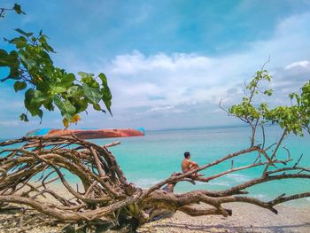 Shirtless man sitting on tree at beach against sky