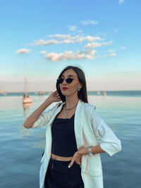 Young woman wearing sunglasses standing at sea against sky