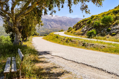 Two parallel mountain roads in crete with a wooden bench in the foreground and snow-capped mountains