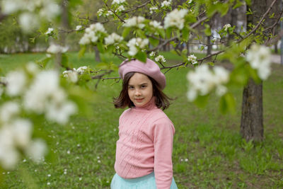 Beautiful little girl stands under the branches of a blossoming white apple tree in the park.