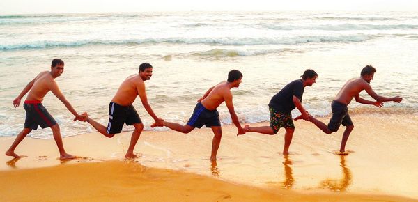 Side view of playful male friends holding legs while standing in row on shore at beach
