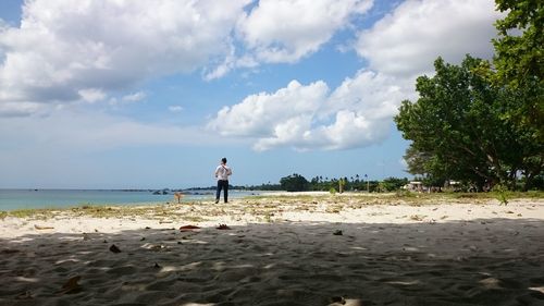 Rear view of man standing on beach against cloudy sky