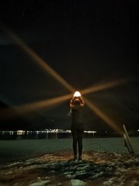 Rear view of man standing by illuminated light against sky at night