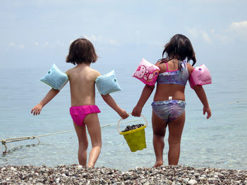 Rear view of sisters holding bucket at beach against sky
