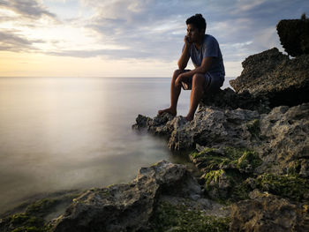 Woman sitting on rock looking at sea shore against sky