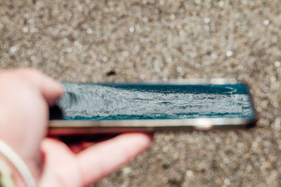 Close-up of human hand holding smart phone reflected sea against sand