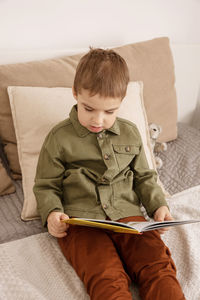 Little and cute caucasian boy reading a book on the bed at home. i