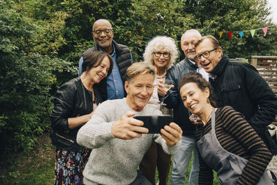 Smiling senior man taking selfie with female and male friends during garden party at back yard