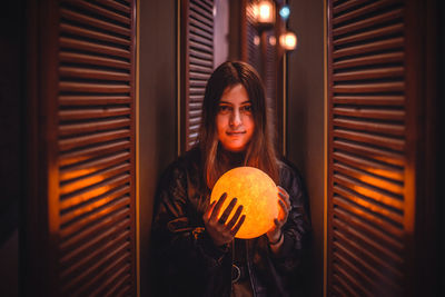 Young woman holding pumpkin while standing against illuminated wall