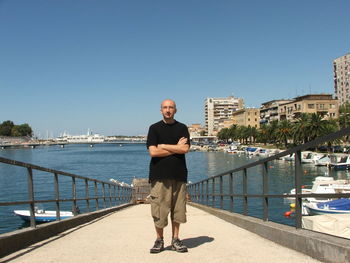Full length of man standing with arms crossed on pier by city against clear blue sky
