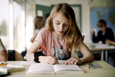 Female student writing in book while sitting at table in classroom