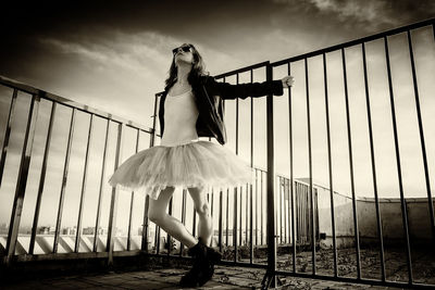 A ballerina in tutu stands at the fence on the roof at sunset in boots and a jacket