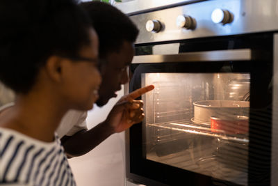 Black couple baking cake for special occasion together at home, looking inside oven with impatience