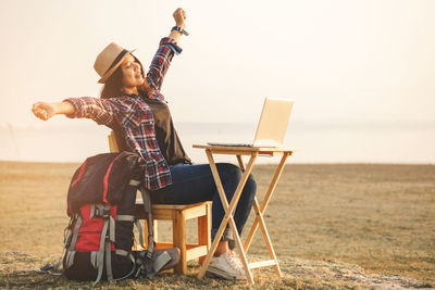 Full length of young man sitting on chair at sea shore against sky