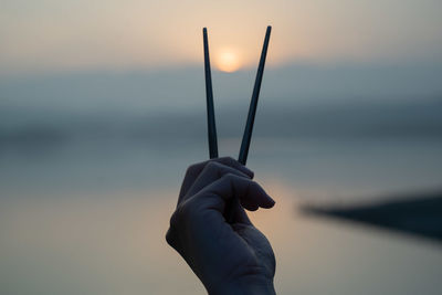 Close-up of person holding chopsticks against sky during sunset