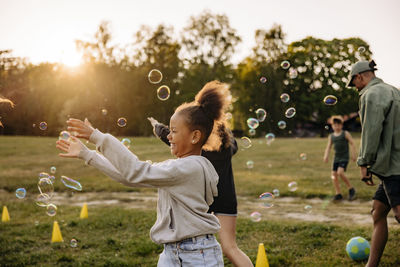 Side view of happy girl catching bubbles while playing with friends in playground at summer camp