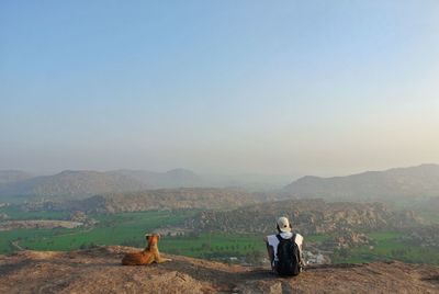 Rear view of man sitting on cliff against sky