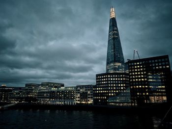 Illuminated buildings by river against cloudy sky