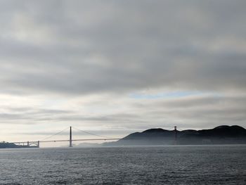 View of suspension bridge over sea against cloudy sky